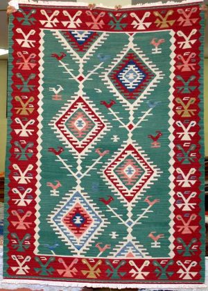 Tappeto contemporaneo kilim lana con motivi decorativi a rombo uncinati su fondo verde e uccelli stilizzati. Spesso bordo rosso decorato con motivi decorativi astratti e lineari